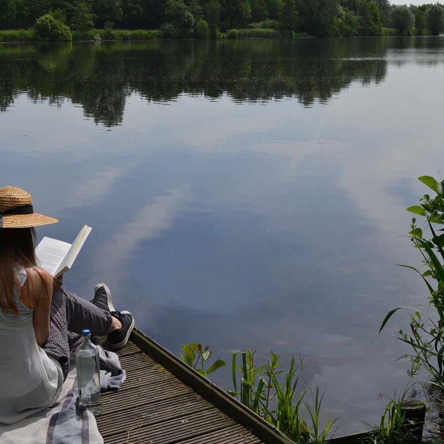Frau mit Hut liest Buch auf Steg am Wasser