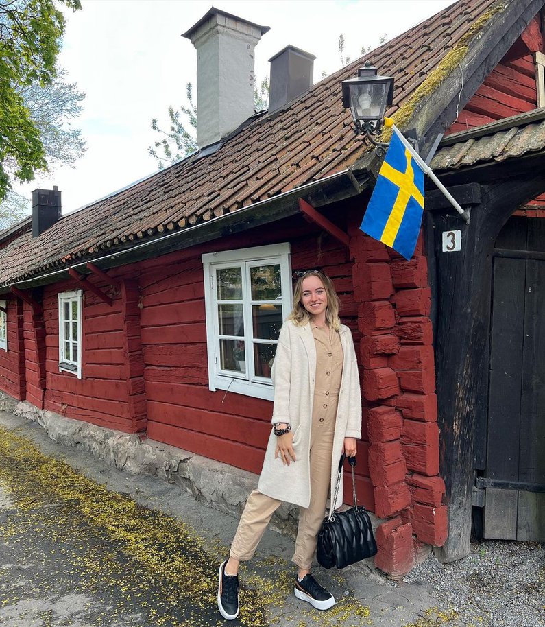 Emely steht vor einem typosch rot gestrichenen Holzhaus, an dem eine blau gelbe schwedische Flagge aufgehängt ist