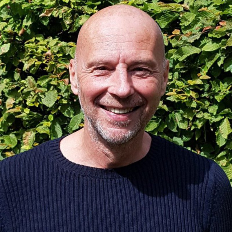 Ein Mann mit schwarzem Pullover posiert vor einer grünen Hecke und lächelt in die Kamera.
