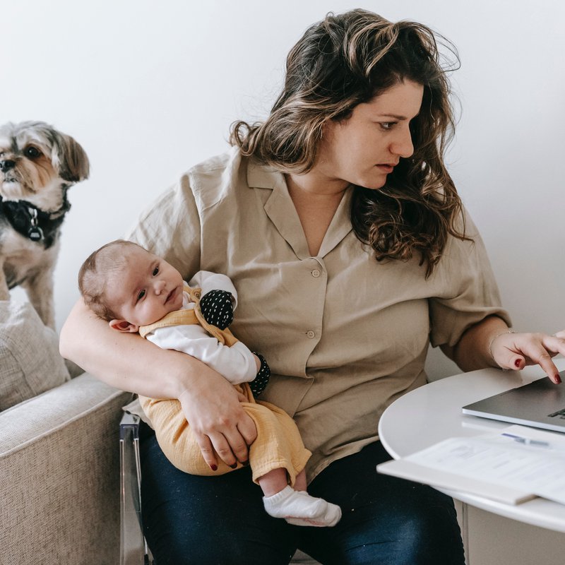 Frau mit Kind im Arm sitzt am Tisch und schaut auf einen Laptop vor sich