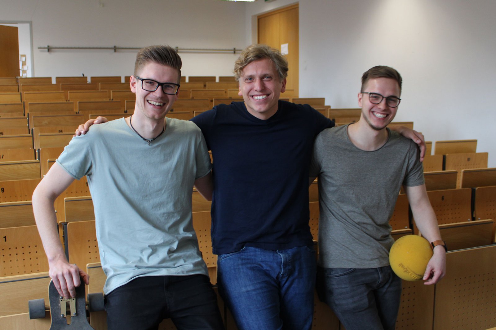 Drei Männer posieren lächelnd in einem leeren Hörsaal.