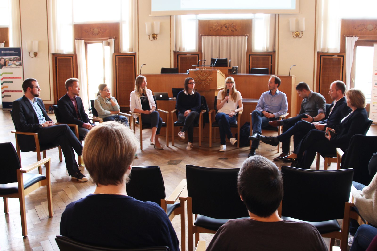 Ein Stuhlkreis in dem eine gemischte Gruppe von Menschen sitzt und diskutiert. Der Raum enthält sehr viele hölzerne Elemente.