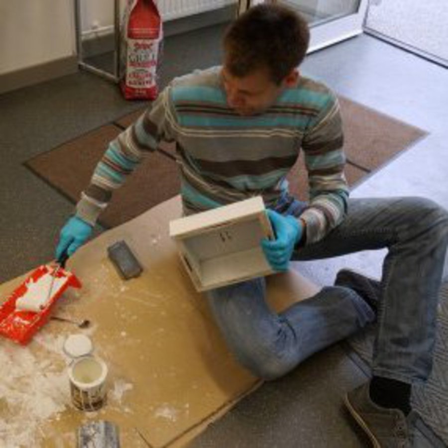 Ein Mann sitzt auf einer Werkplatte und streicht einen Rahmen.