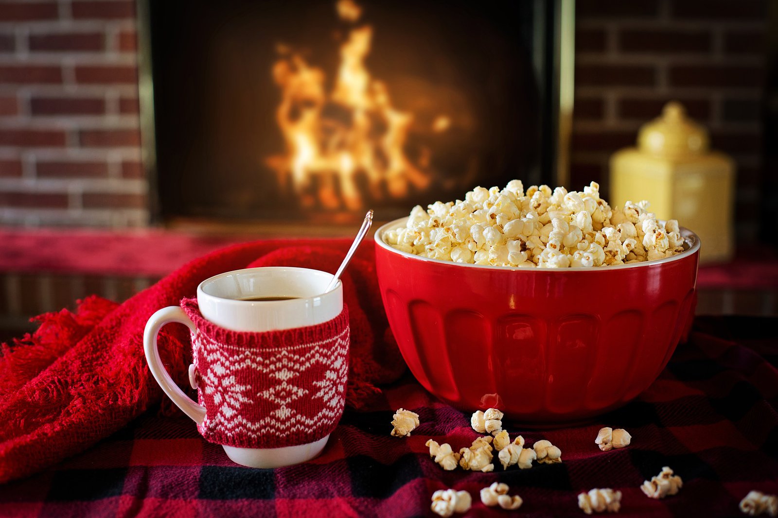 Eine weihnachtliche Tasse und eine rote Schale mit Popcorn, im Hintergrund lodert eine Flamme, in einem Kamin.