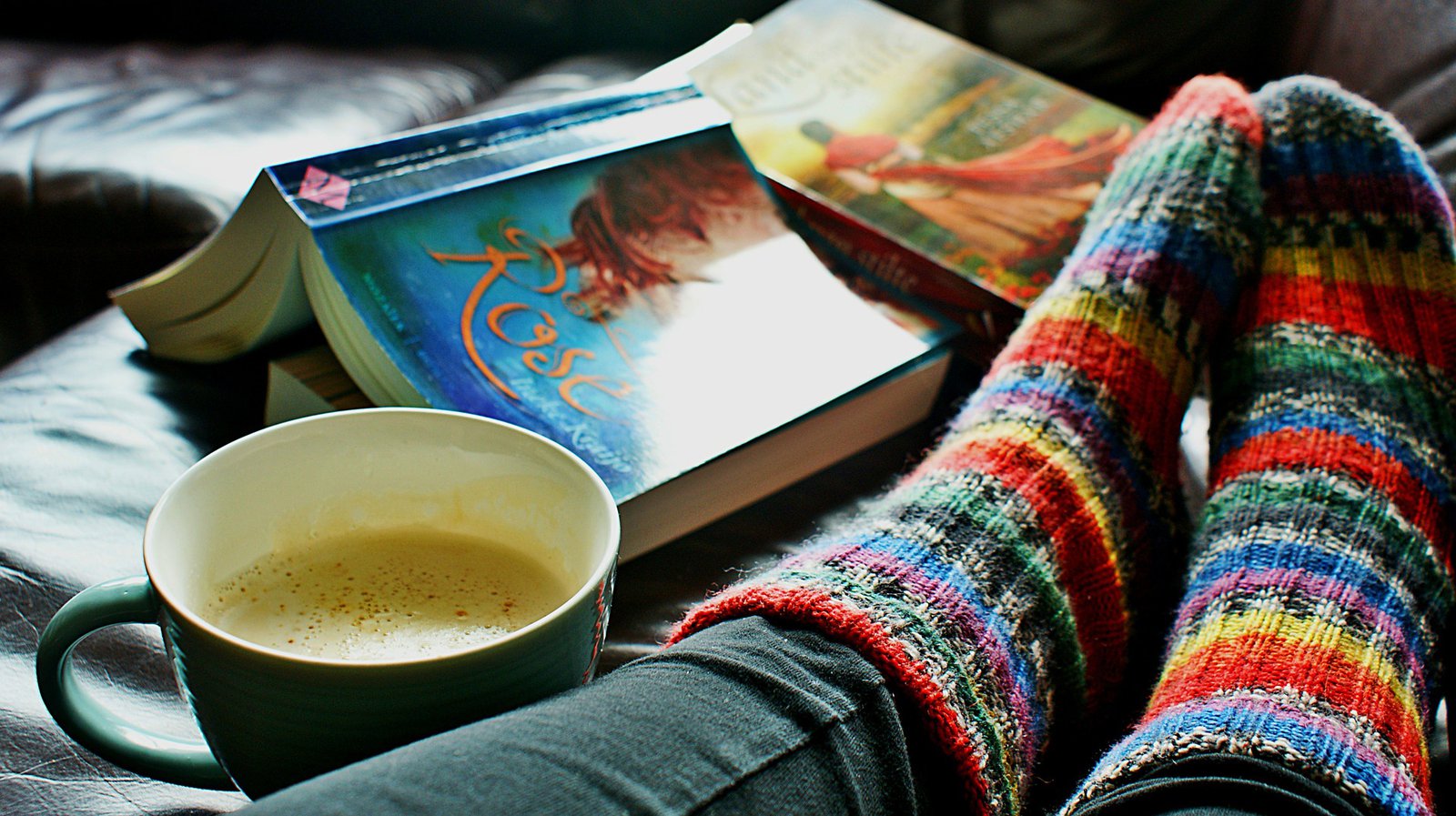 Mit bunten, gestrickten SOcken bekleidete Füße. Drumherum eine Kaffeetasse und zwei Bücher.