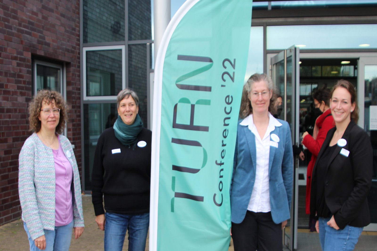 Vier Frauen schauen lächelnd in die Kamera, zwischen Ihnen steht eine Flagge. Auf dieser steht "TURN Conference '22" geschrieben.