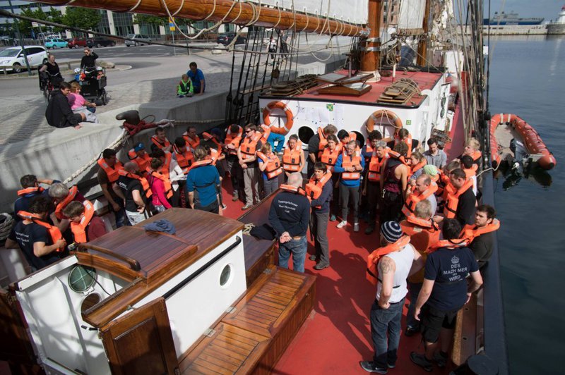 Eine Gruppe Studierender befindet sich auf einem Segelschiff. Alle sind mit einer orangenen Rettungsweste ausgestattet.