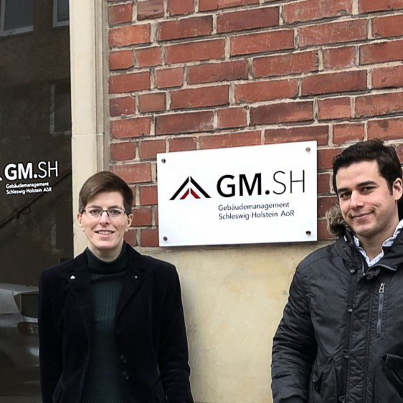 Ein Mann und eine Frau posieren vor einer hohen Glastür mit massivem Holzrahmen. Auf dem Schild neben dem sie stehen steht "GM.SH".