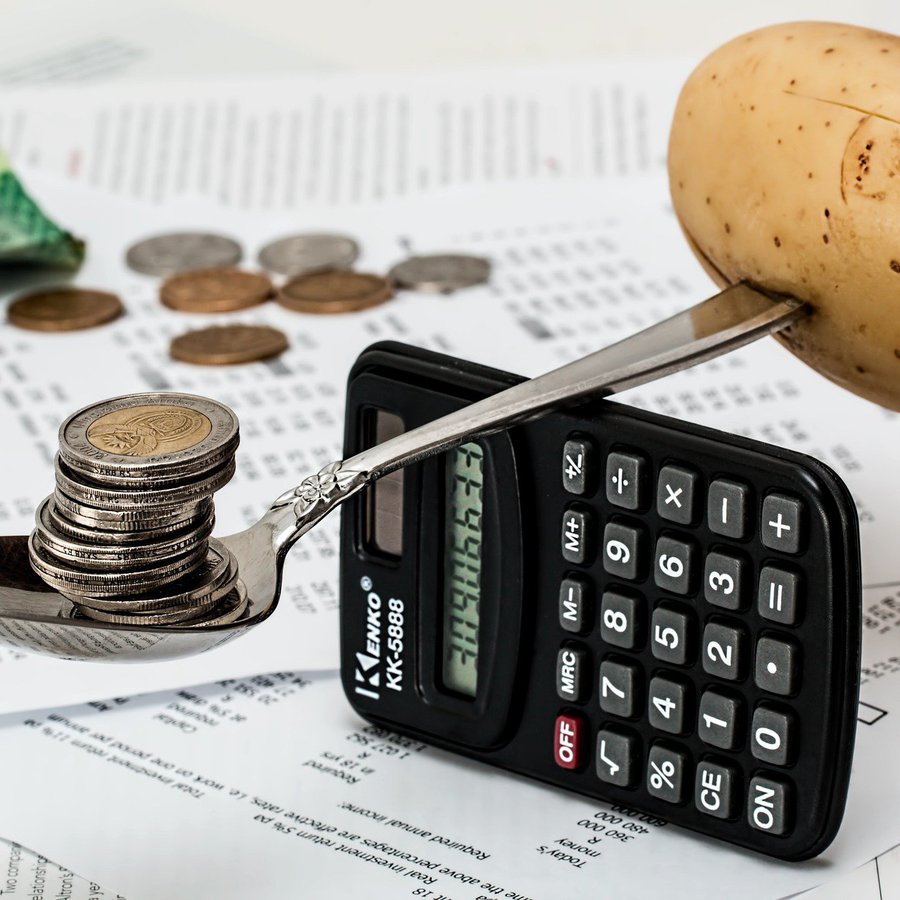 Taschenrechner hochkant mit Löffel darauf, der Balance hält zwischen Münzen und Kartoffel