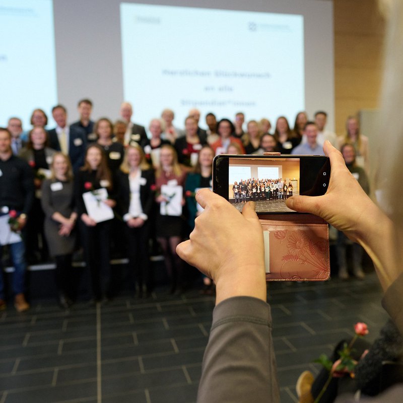 Eine Frau wird von hinten gezeigt, die mit ihrem Smartphone die Stipendita*innen auf der Bühne des Audimax fotografiert. 