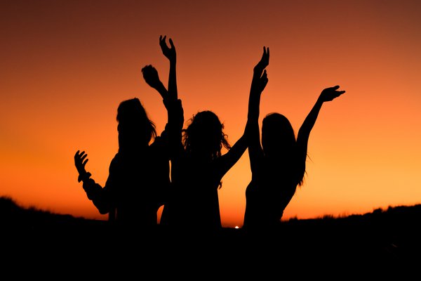Schwarze Silhouetten dreier Frauen die im Sonnenuntergang tanzen.