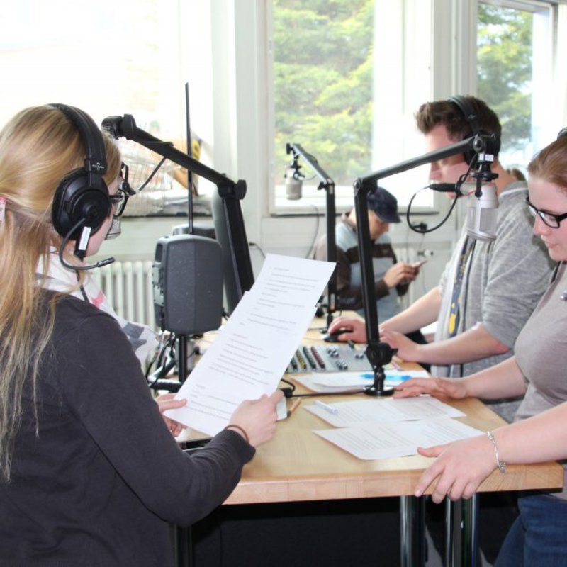 Zwei Frauen und ein Mann stehen mit Kopfhörern ausgestattet vor ihren Mikrofonen.