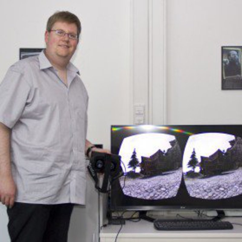 Ein Mann in grauem Kurzarmhemd, steht neben einem Monitor.