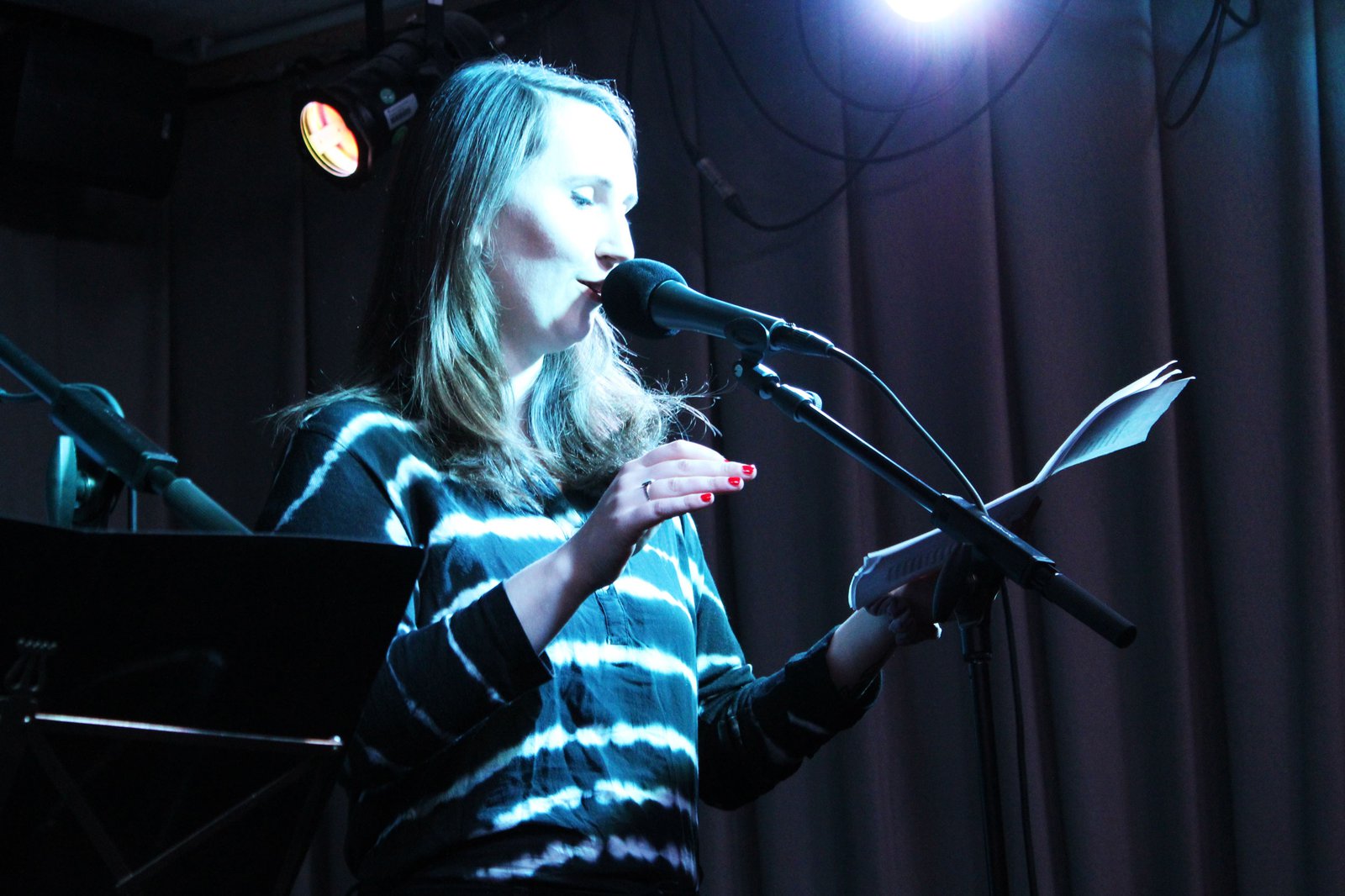Eine Frau steht vor einem Mikrofon, auf einer schwach beleuchteten Bühne und liest etwas aus dem Skript in ihrer Hand ab.