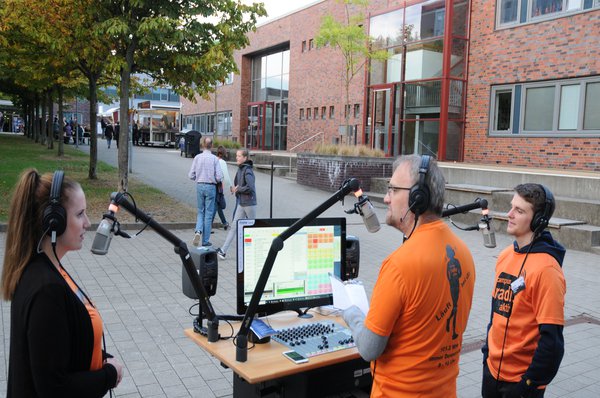Zwei Männer und eine Frau stehen, in orangenen Shirts, um ein im Freien aufgebautes Radiopult.