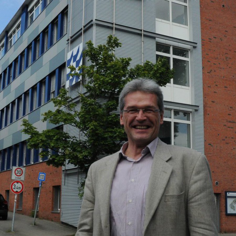 Ein Mann in hellem Sakko steht vor einem Universitätsgebäude und lächelt freudig in die Kamera.