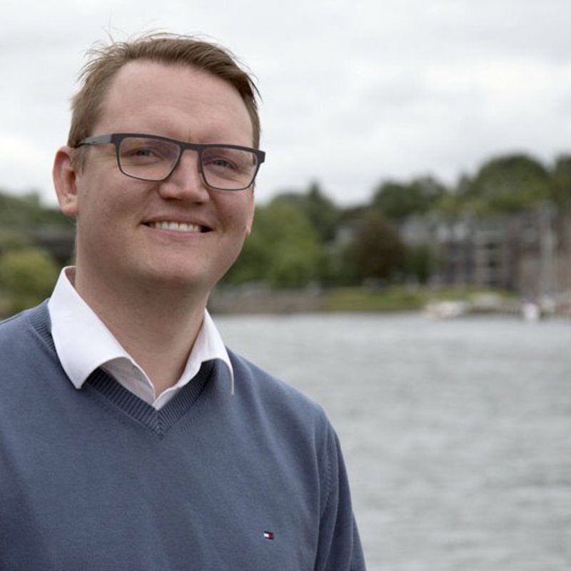 Ein Mann mit blauer Brille und blauem Pullover posiert, nahe der Schwentinemündung, für die Kamera.