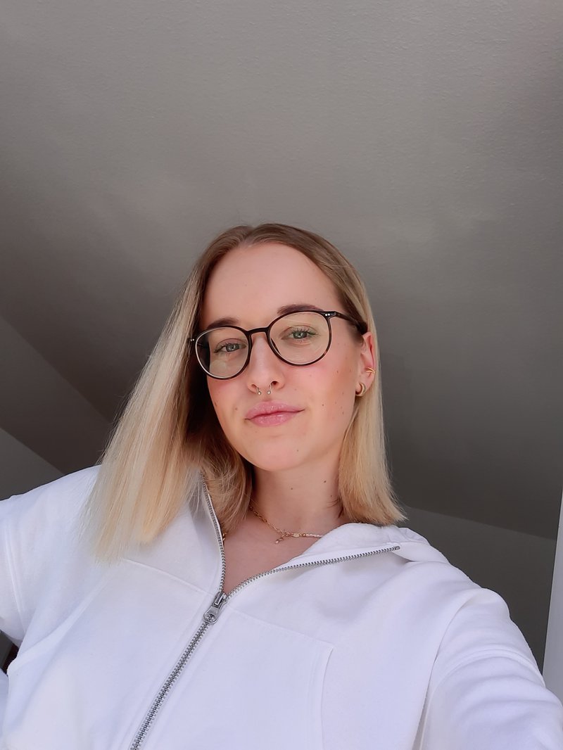 Ein Selfie von Emely, sie trägt eine Brille, hat blonde schulterlange Haare und eine weiße Jacke an