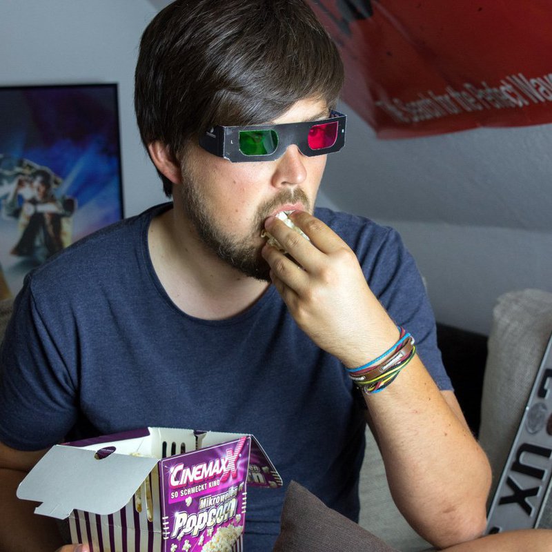Ein Mann mit 3D-Brille sitzt in seinem Zimmer und isst Popcorn.