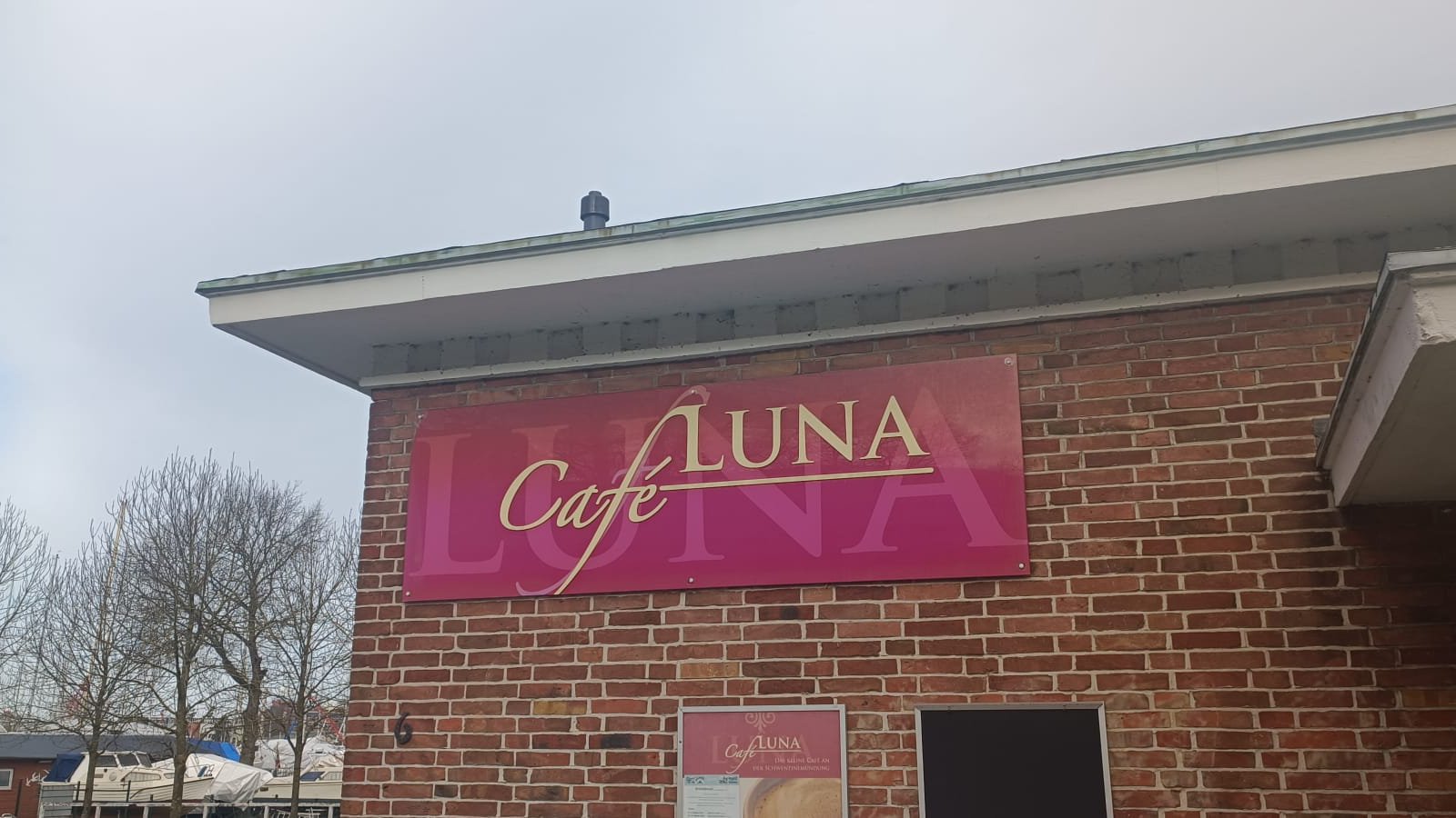 Das rote Ladenschild vom Café Luna hängt an der Hausecke. Im Hintergrund sieht man grauen Himmel.