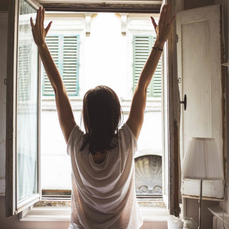 Eine Frau streckt ihre Arme, vor einem geöffneten Fenster.