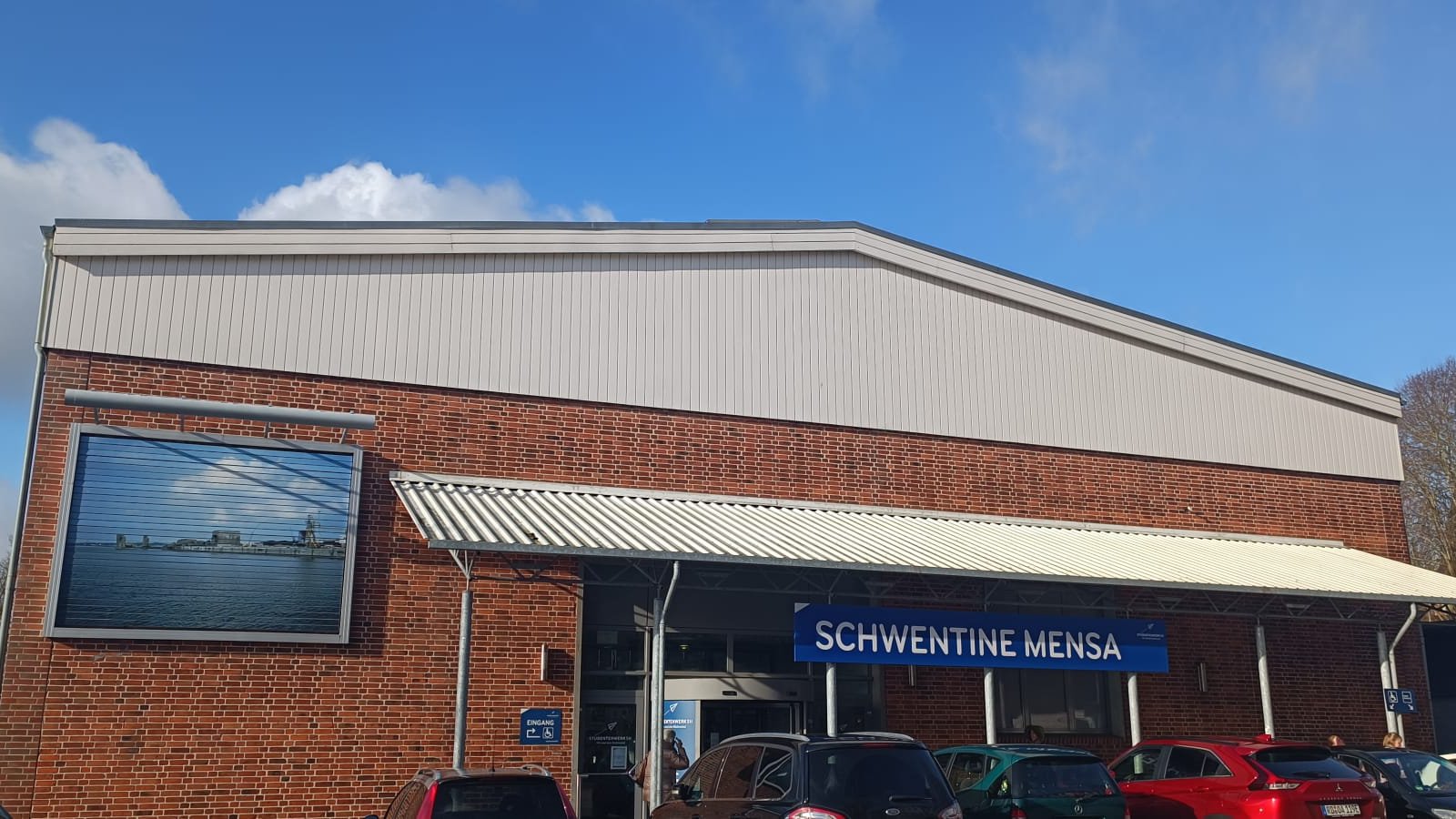 Ein Backsteingebäude mit der Aufschrift "Schwentine-Mensa". Im Vordergrund parken Autos, im Hintergrund ein beinahe wolkenloser Himmel.