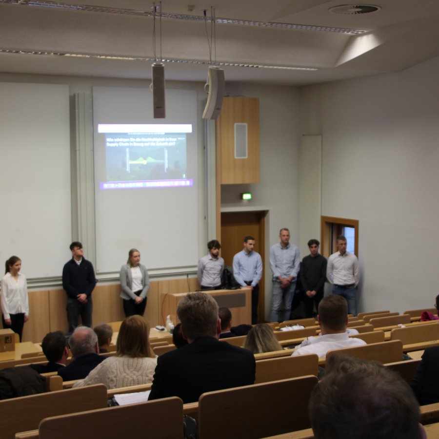 Neun Studierende stehen vor einem Publikum in einem Hörsaal. Der Professor gestikuliert.