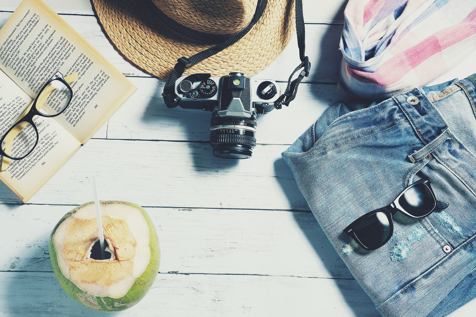 Eine Trinkkokosnuss, ein Buch und eine Brille, ein Hut, eine Kamera, ein Schal, eine Jeans und eine Sonnenbrille liegen auf einer Picknickbank.