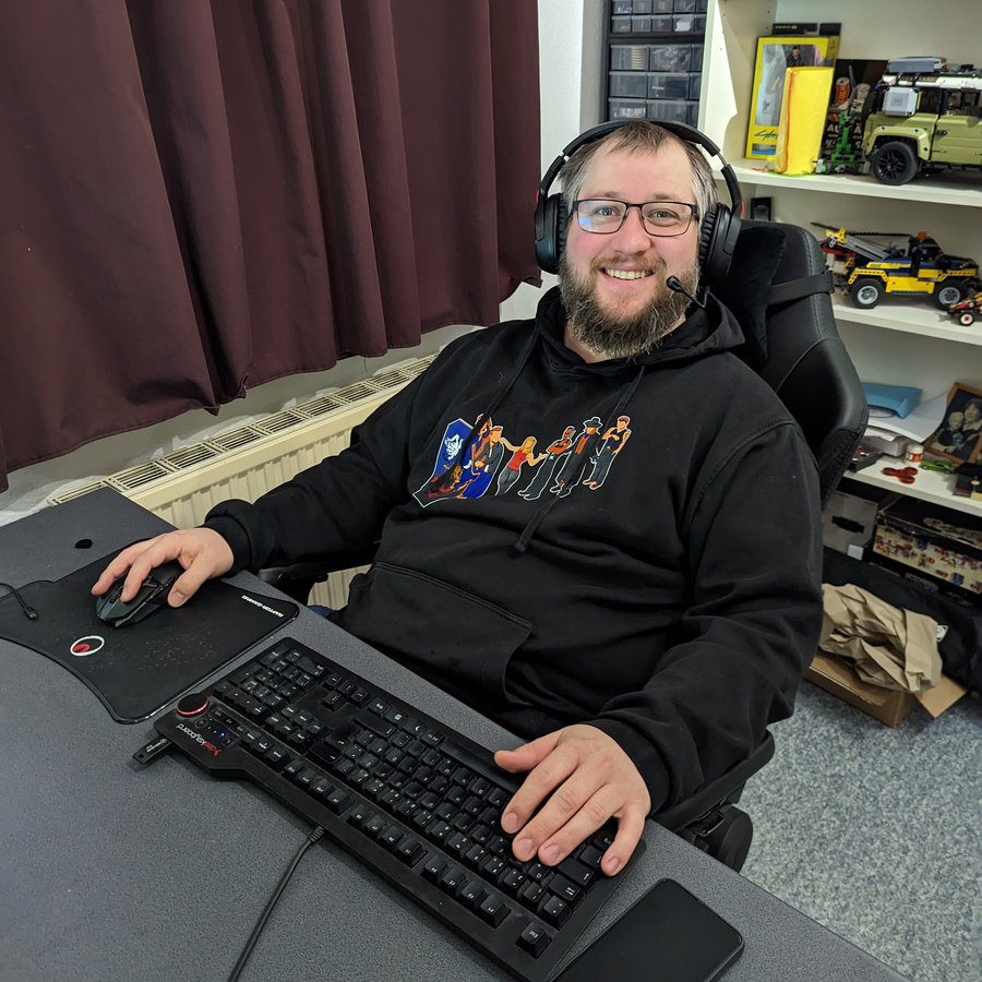 Ein Mann sitzt in einem Bürostuhl am Computer, trägt ein Headset, hat eine Hand auf der Tastatur und eine an der Computermaus und schaut lächelnd in die Kamera
