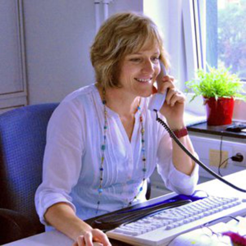 Eine Frau in weißer Bluse sitzt beschäftigt, aber glücklich, an ihrem Arbeitsplatz. In der einen Hand hält sie das Telefon, in der anderen die Maus.