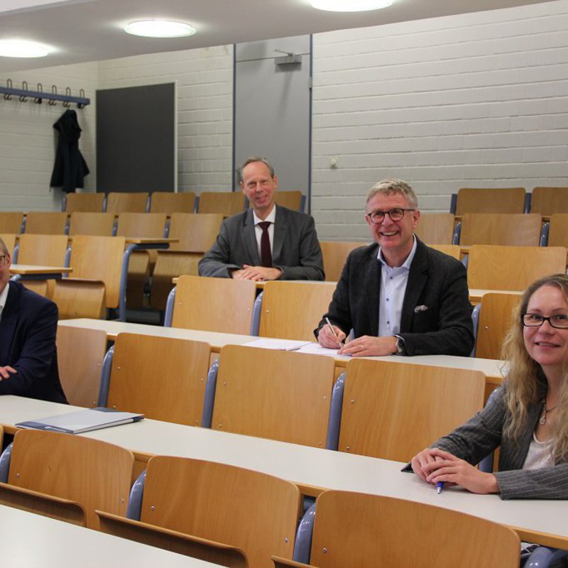 In einem Hörsaal sitzen links vorne Prof. Dr. Björn Christensen, vorne rechts Katja Buhs, dahinter Dr. Edgar Schmitt und eine Reihe dahinter Prof, Dr. Ulrich Jetzek.