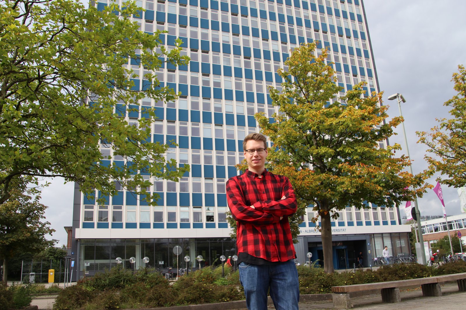 Ein Student in einem schwarz-rot kariertem Hemd steht vor einem hohen Gebäude auf dem Campus.