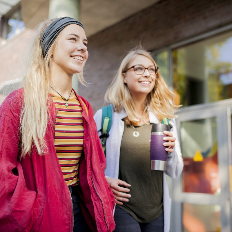Zwei junge Frauen gehen eine Straße entlang, eine von beiden hält einen To-Go-Becher in der Hand.