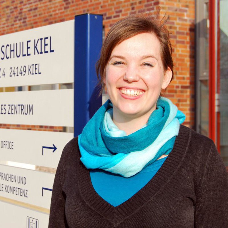 Eine Frau mit blauem Schal, lächelt freundlich vor dem International Office der FH Kiel.