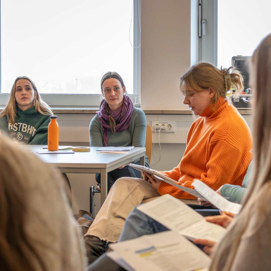 Eine Frau in einem organenen Pullover sitzt auf einem Stuhl und blickt auf einen Schreiblock auf ihrem Schoß. Im Hintergrund sitzen zwei Frauen, die ihr zuhören.