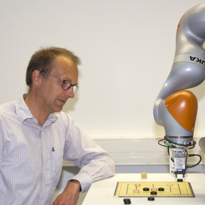 Ein Mann in wieß-kariertem Hemd spielt Mühle gegen einen Roboter.