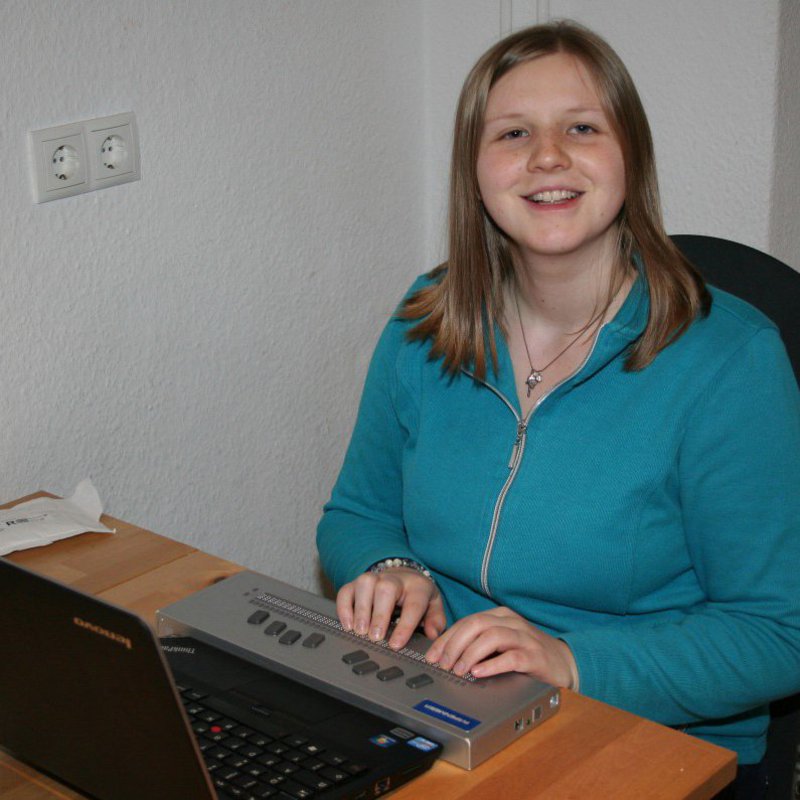 Eine Frau in grünem Pullover sitzt vor ihrem Laptop und berührt mit den Händen, ein davor geschaltetes Gerät.