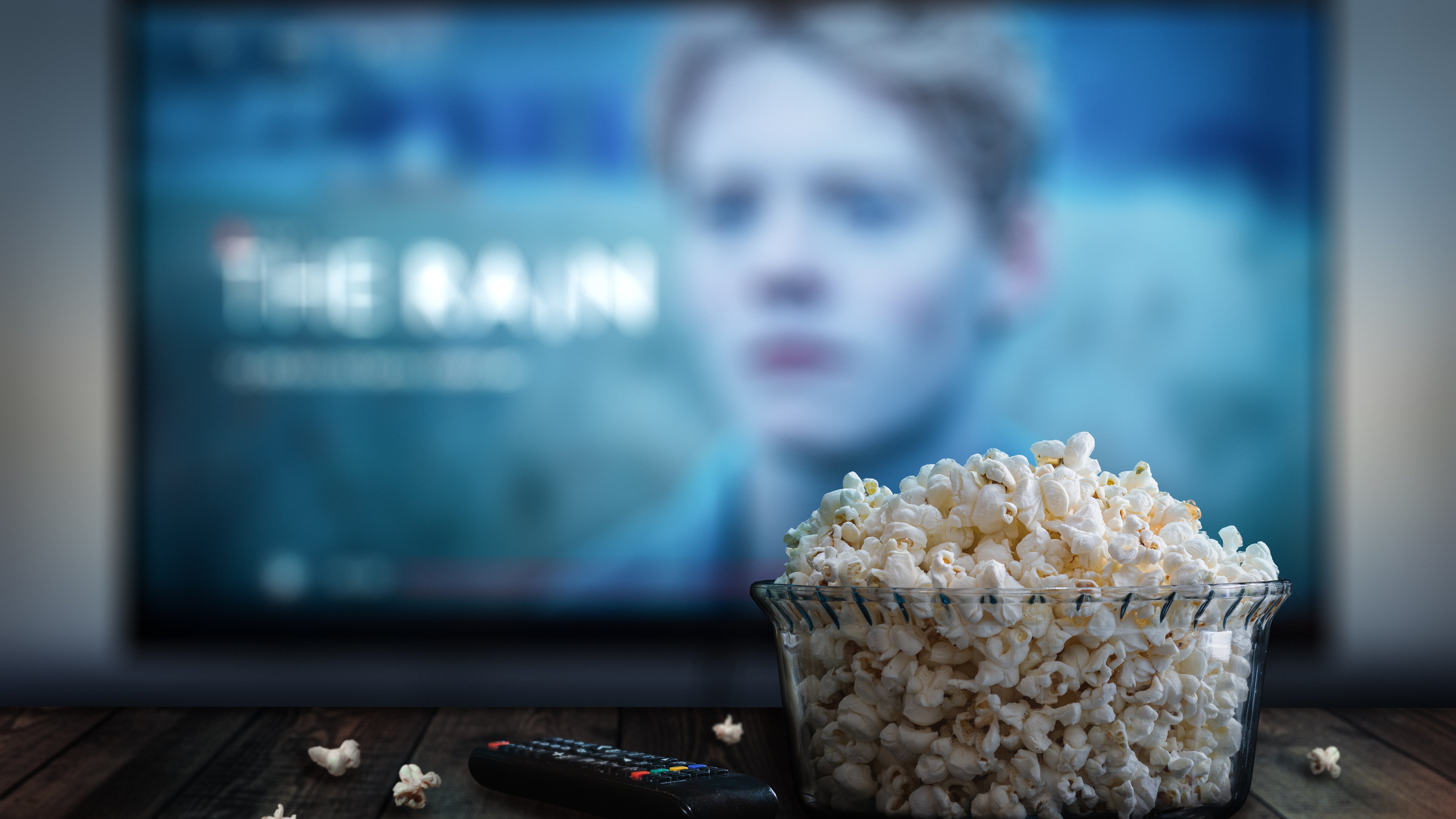 eine Schale mit Popcorn steht auf einem Tisch neben einer Fernbedienung, dahinter verschwommen ein TV-Bildschirm