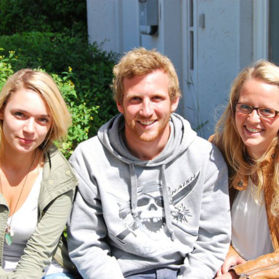 Zwei Frauen und eine Mann sitzen zusammen, bei Sonnenschein, auf einer Bank im Grünen.