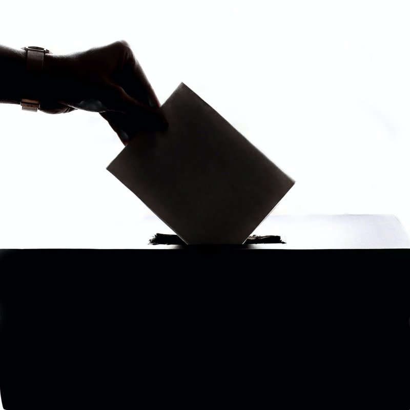 Zu sehen ist eine Hand, die einen Briefumschlag hält und in einen Karton (=Wahlurne) steckt. 
