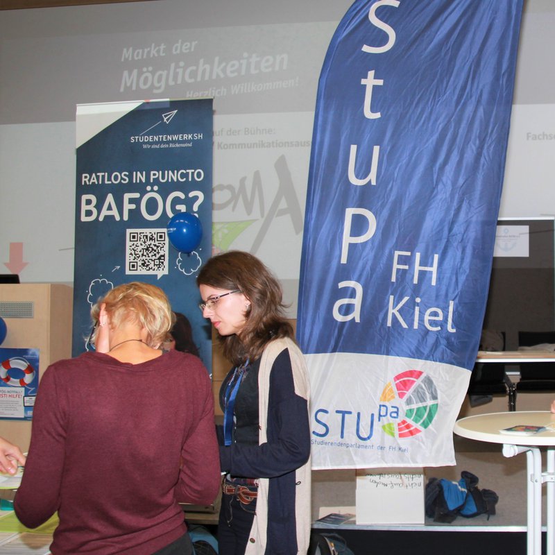 2 Studentinnen und 2 Studenten stehen in einem größeren Raum an Stehtischen, es ist eine Beachflag mit der Aufschrift StuPa zu sehen und ein Aufsteller vom Studentenwerk, die für Bafög-Beratung wirbt.