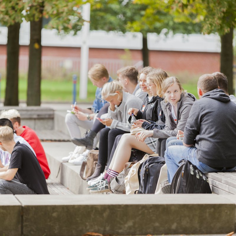 Eine Gruppe Studierender sitzt auf dem Campus, die Sonne scheint. 