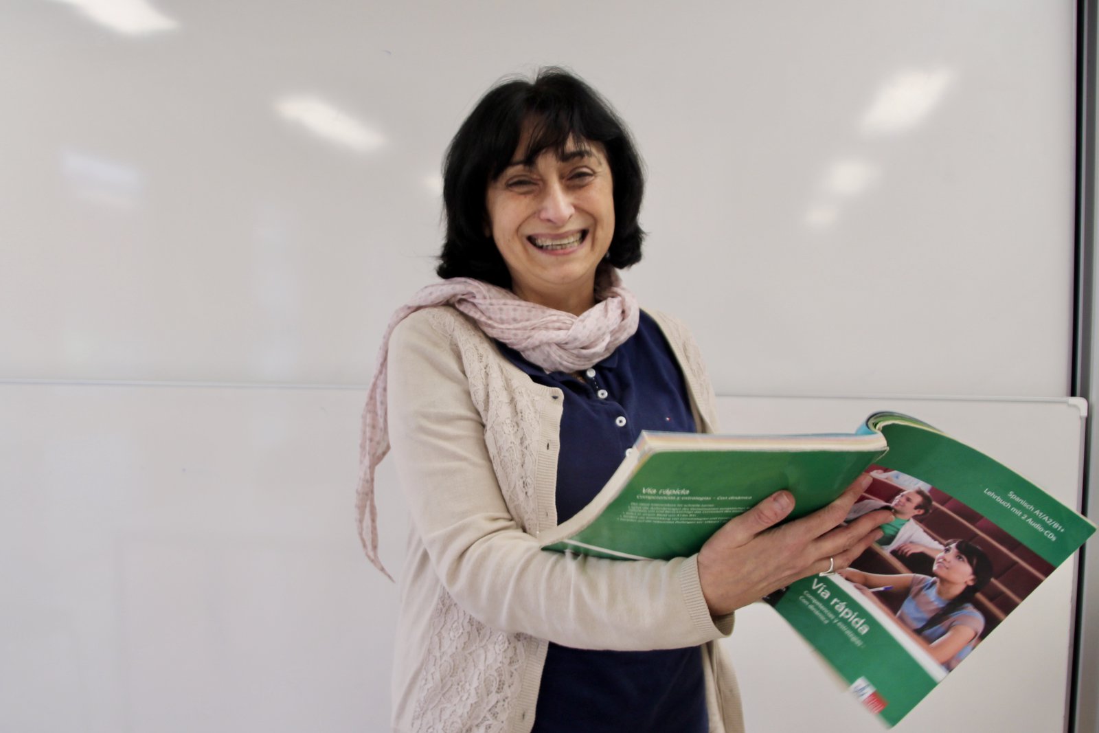 Eine breit lächelnde Frau, mit einem großen, grünen Lehrbuch in der Hand.