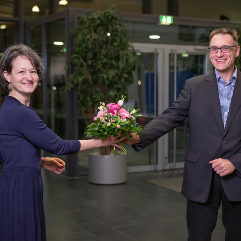 Dr. Anja Franke-Schwenk steht im Foyer des Mehrzweckgebäudes. Prof. Dr. Claus Neumann gratuliert ihr zur Wahl zur Kanzlerin mit einem Blumenstrauß.