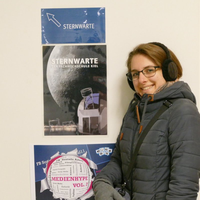 Eine Frau mit Winterjacke und Ohrenwärmern posiert vor Plakaten der Sternenwarte Fachhochschule Kiel.
