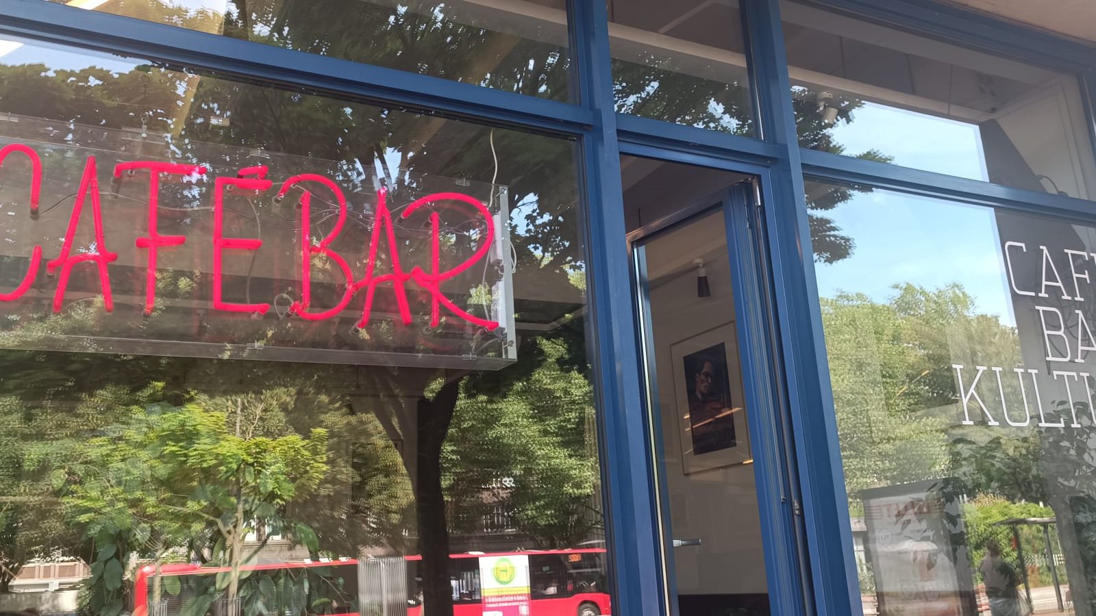 Ein Fenster, in welchem sich Bäume spiegeln. Eine Anzeige aus Neon-Röhren schreibt "Cafébar" auf die Scheibe.