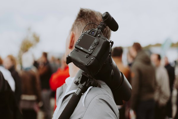Ein Mann trägt eine Kamera mit stativ über der Schulter und wendet seinen Blick auf die hinter ihm stehende Menge.