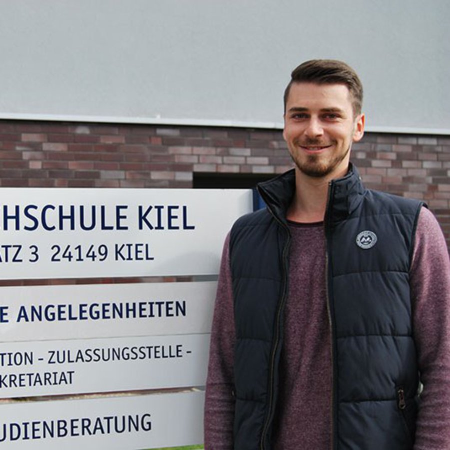 Ein Mann steht neben dem Eingangsschild  des Referats für Studentische Angelegenheiten, der FH Kiel..