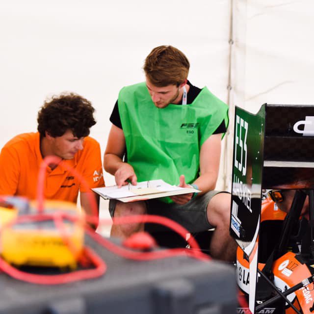 Ein Mann mit orangefarbenem Shirt und ein Mann mit grünem Shirt schauen auf ein Dokument