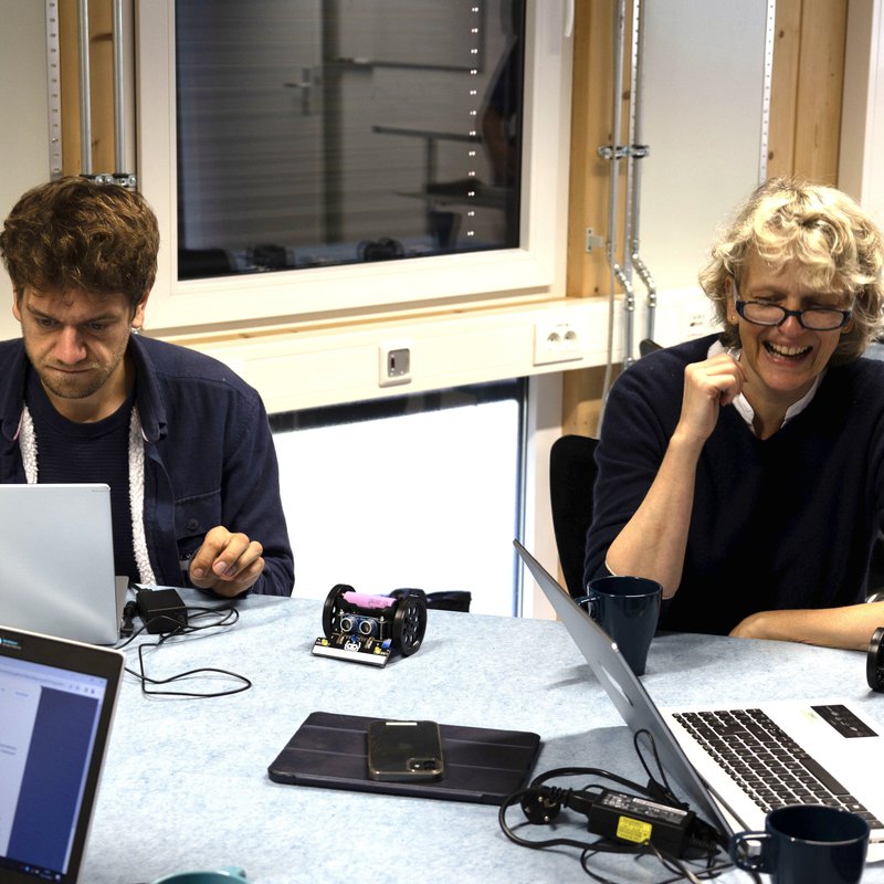 Ein Mann und eien Frau sitzen an Computern, zwischen ihnen steht auf dem Tisch ein etwa handtellergroßer Roboter